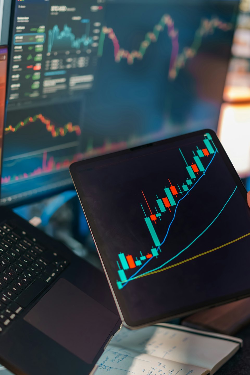 Mano che tiene un tablet con un grafico finanziario sopra un laptop.